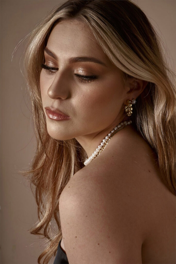 Clarisa Cisneros a model with a radiant complexion showcases a side profile with her eyes closed revealing meticulous makeup and a pearl necklace.